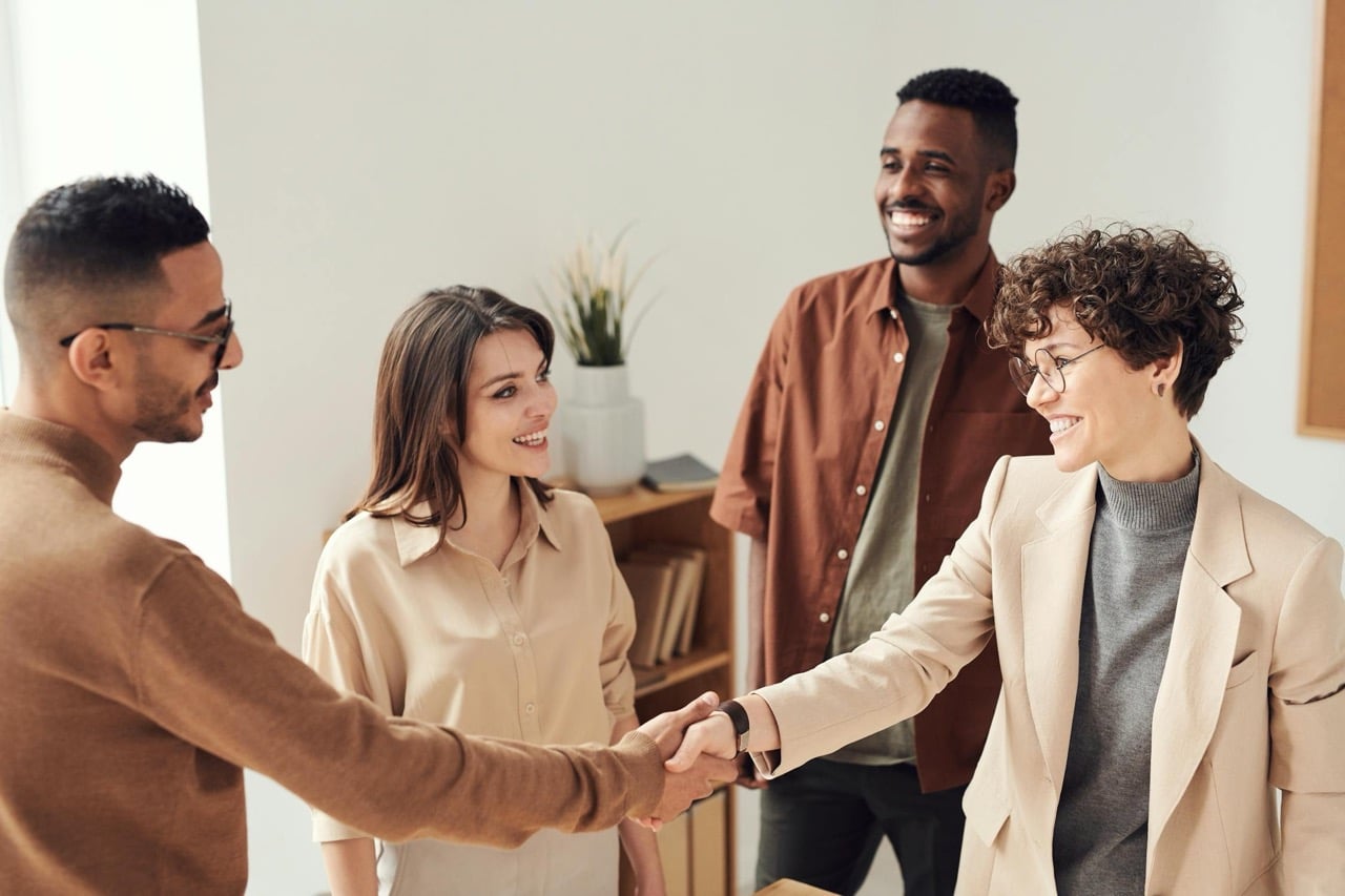 A group of four people are having a friendly meeting in a bright office, showcasing leadership skills. Two are shaking hands, while the others smile and watch. Everybody is casually dressed, and there's a plant in the background.