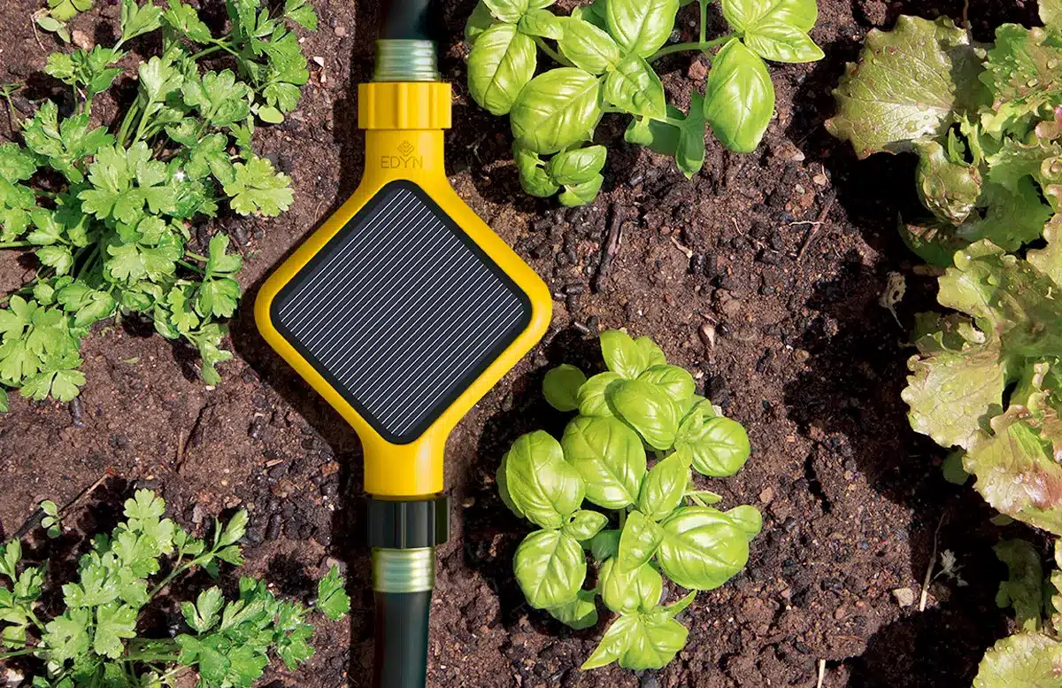 A smart garden sensor with a yellow casing is inserted into the soil among green plants, including basil and lettuce. The device has a solar panel on top and is connected to a garden hose, surrounded by dark, rich soil.