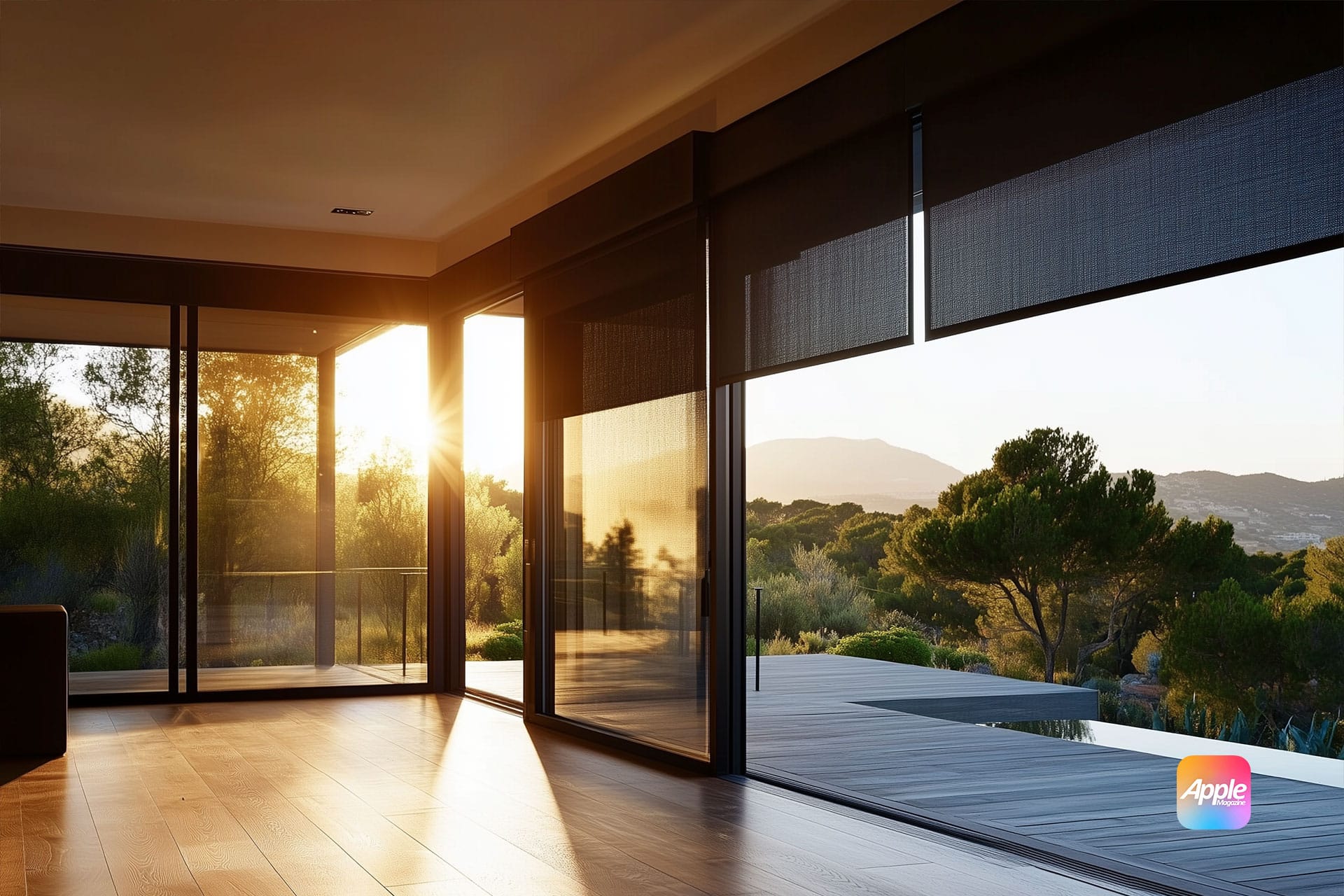 A modern living room boasts large windows with motorized shades, offering a scenic view of a sunset over the landscape. Wooden flooring complements the view of the deck, while a digital Apple logo adorns the corner. Trees and mountains stand serenely in the background.