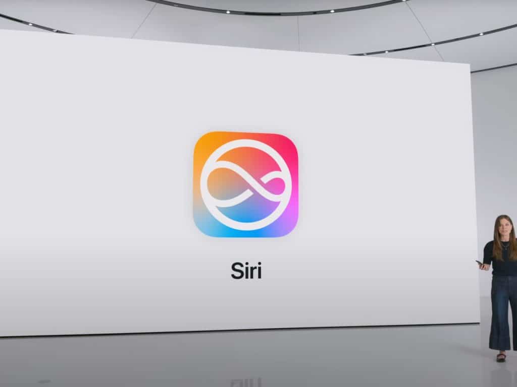 A woman in black clothes stands on a stage presenting a large screen displaying the Siri logo, which features a colorful swirl design. The word "Siri" is shown below the iconic Apple branding. The setting appears modern and minimalist, capturing Apple's sleek aesthetic.