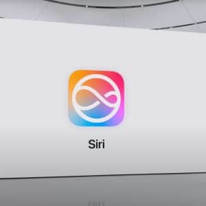 A woman in black clothes stands on a stage presenting a large screen displaying the Siri logo, which features a colorful swirl design. The word "Siri" is shown below the iconic Apple branding. The setting appears modern and minimalist, capturing Apple's sleek aesthetic.