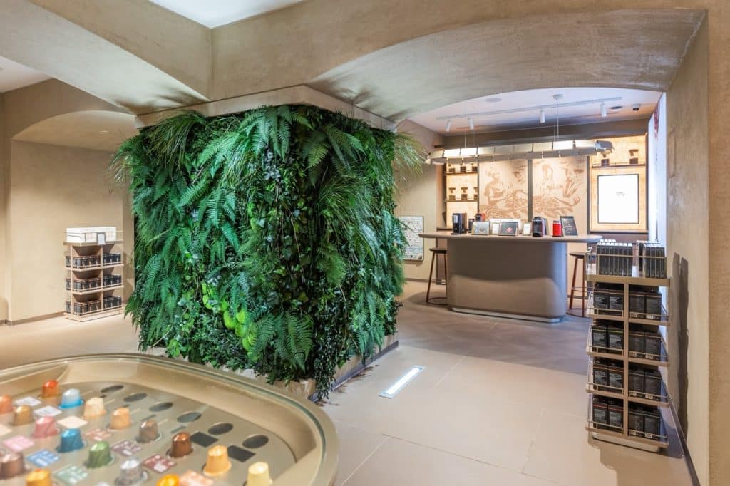 A modern coffee shop interior with a wall covered in green plants. In the foreground, a display with coffee capsules. A counter with bar stools is in the background, and shelves with merchandise are on the right.