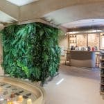 A modern coffee shop interior with a wall covered in green plants. In the foreground, a display with coffee capsules. A counter with bar stools is in the background, and shelves with merchandise are on the right.