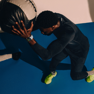 Custom Workouts - A person wearing black athletic gear and neon green shoes is exercising on a blue floor, holding a large medicine ball. Glancing at their Apple Watch Series 10 for metrics, the iconic Apple logo gleams in the corner, merging tech with fitness seamlessly.