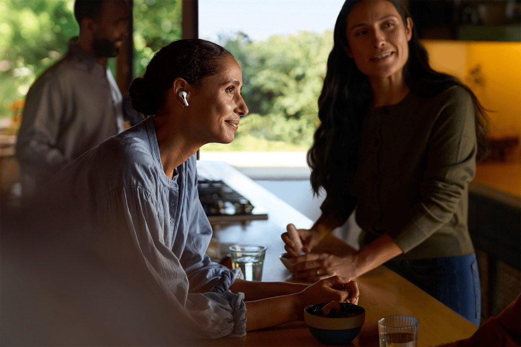 Two women are in a kitchen, one wearing wireless earbuds for health monitoring and leaning on a counter, while the other holds a bowl and spoon. A man stands by a window in the background. The counter is adorned with glasses of water and a bowl of fruit.