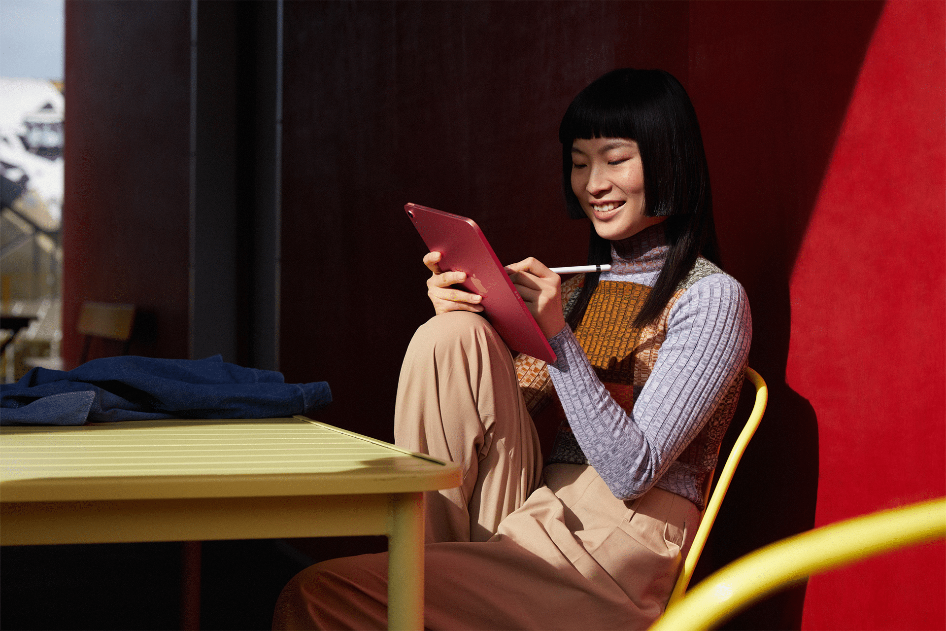 A person sits at a yellow table against a red wall, smiling while using an iPad 11th Generation with a stylus. They are wearing a patterned sweater and beige pants, with a folded blue jacket on the table. Sunlight creates contrasting shadows.