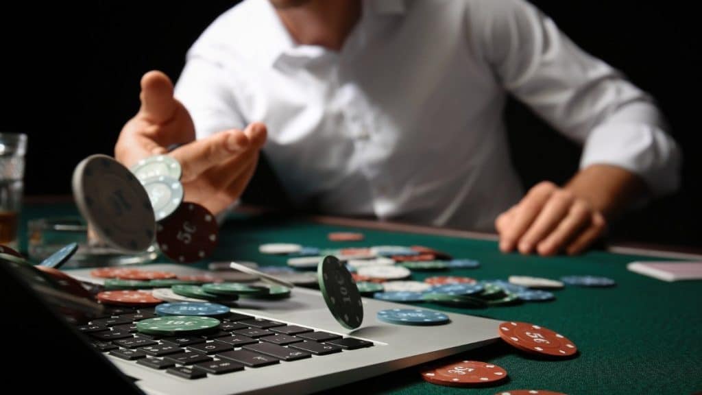 A person in a white shirt throws poker chips onto a green poker table, with some chips scattered on an Apple laptop keyboard. The scene captures the innovative blend of traditional poker gaming and mobile casino gaming technology.