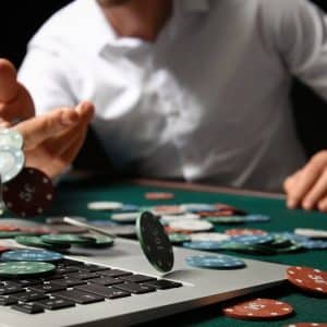 A person in a white shirt throws poker chips onto a green poker table, with some chips scattered on an Apple laptop keyboard. The scene captures the innovative blend of traditional poker gaming and mobile casino gaming technology.