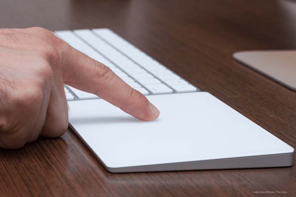 A person using a Magic Trackpad on a desk. The finger is pressing down on the trackpad, with a keyboard visible in the background. The wooden surface of the desk adds a touch of elegance to the scene.