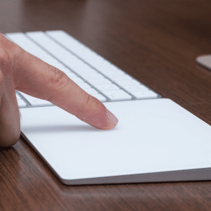 A person using a Magic Trackpad on a desk. The finger is pressing down on the trackpad, with a keyboard visible in the background. The wooden surface of the desk adds a touch of elegance to the scene.