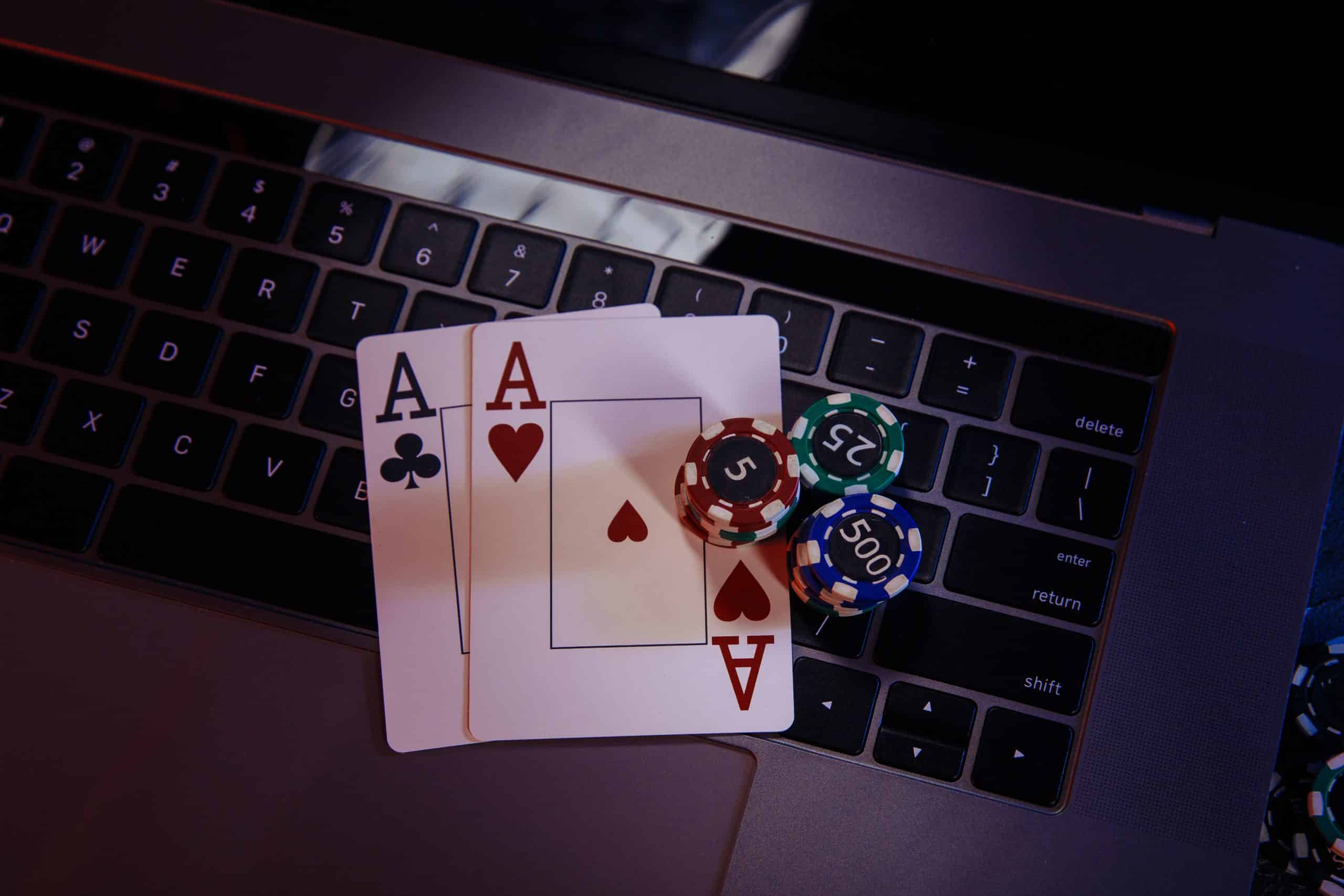 Two Aces of Clubs and Hearts on a laptop keyboard surrounded by poker chips in various denominations and colors, including green 25, purple 500, and red 5.