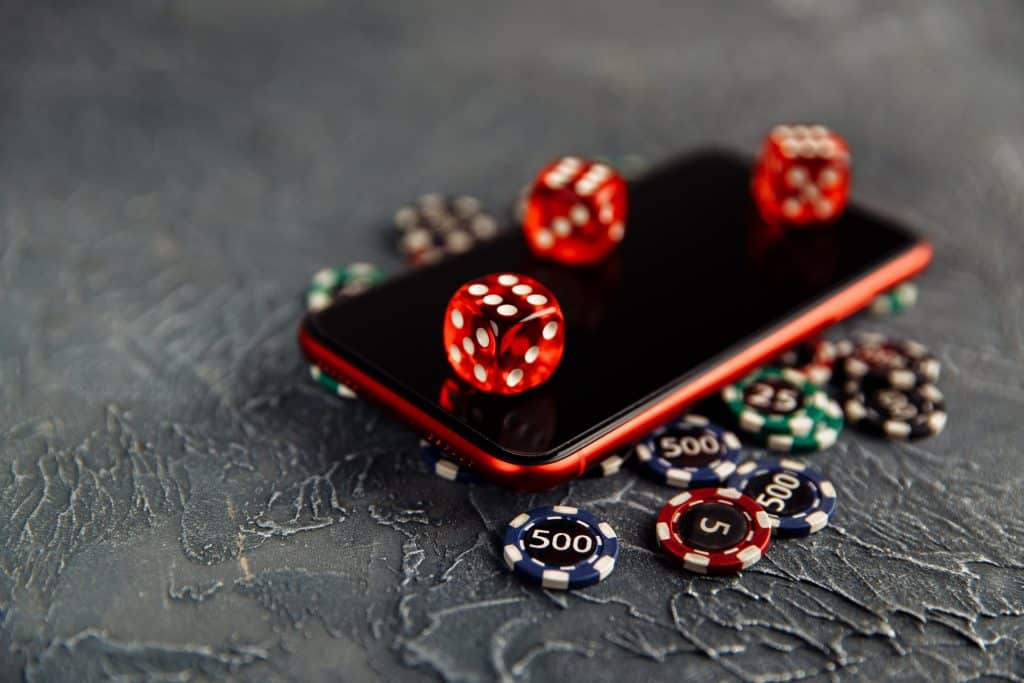 A smartphone lies on a textured surface surrounded by red dice and various poker chips. The phone screen is reflective, showing the bottom side of a red die resting on top. The poker chips display different values.