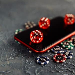 A smartphone lies on a textured surface surrounded by red dice and various poker chips. The phone screen is reflective, showing the bottom side of a red die resting on top. The poker chips display different values.