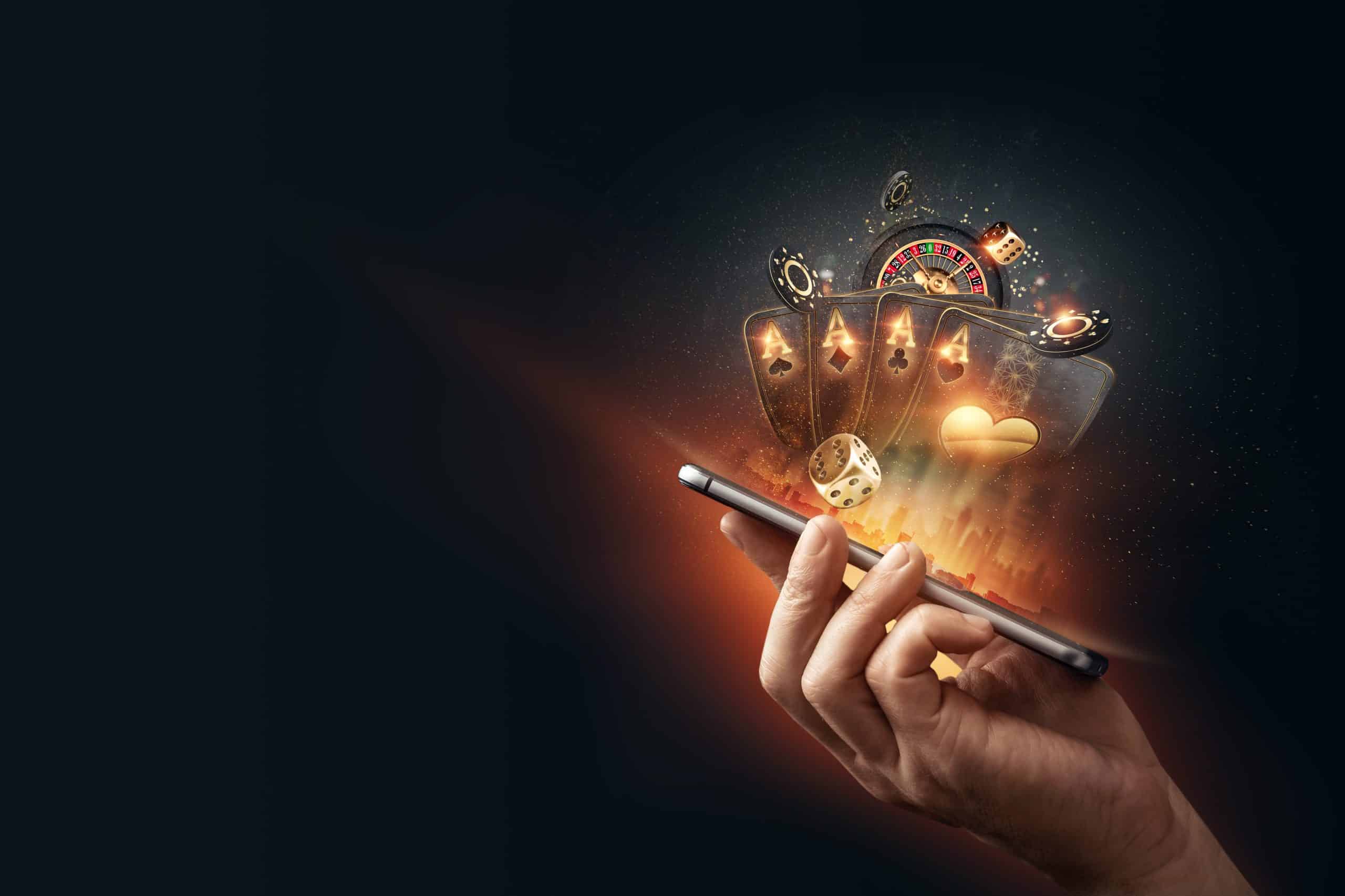 A hand holds a smartphone emitting glowing playing cards, dice, roulette wheel, and poker chips, symbolizing online gambling. The background is dark, highlighting the vivid colors of the gaming elements.