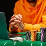 A person in a yellow hoodie sits at a table with a laptop, stacks of poker chips, and a spread of playing cards showing an ace of hearts. Their hands are folded in front of them, focusing intently.