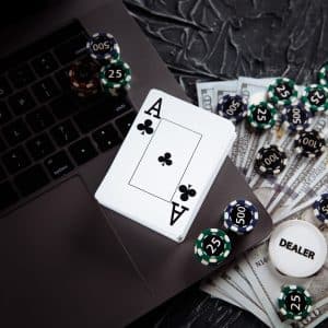 A laptop keyboard surrounded by casino chips, dollar bills, and playing cards, with an Ace of Clubs on top. A dealer button is also visible, suggesting an online gambling or poker game setting.