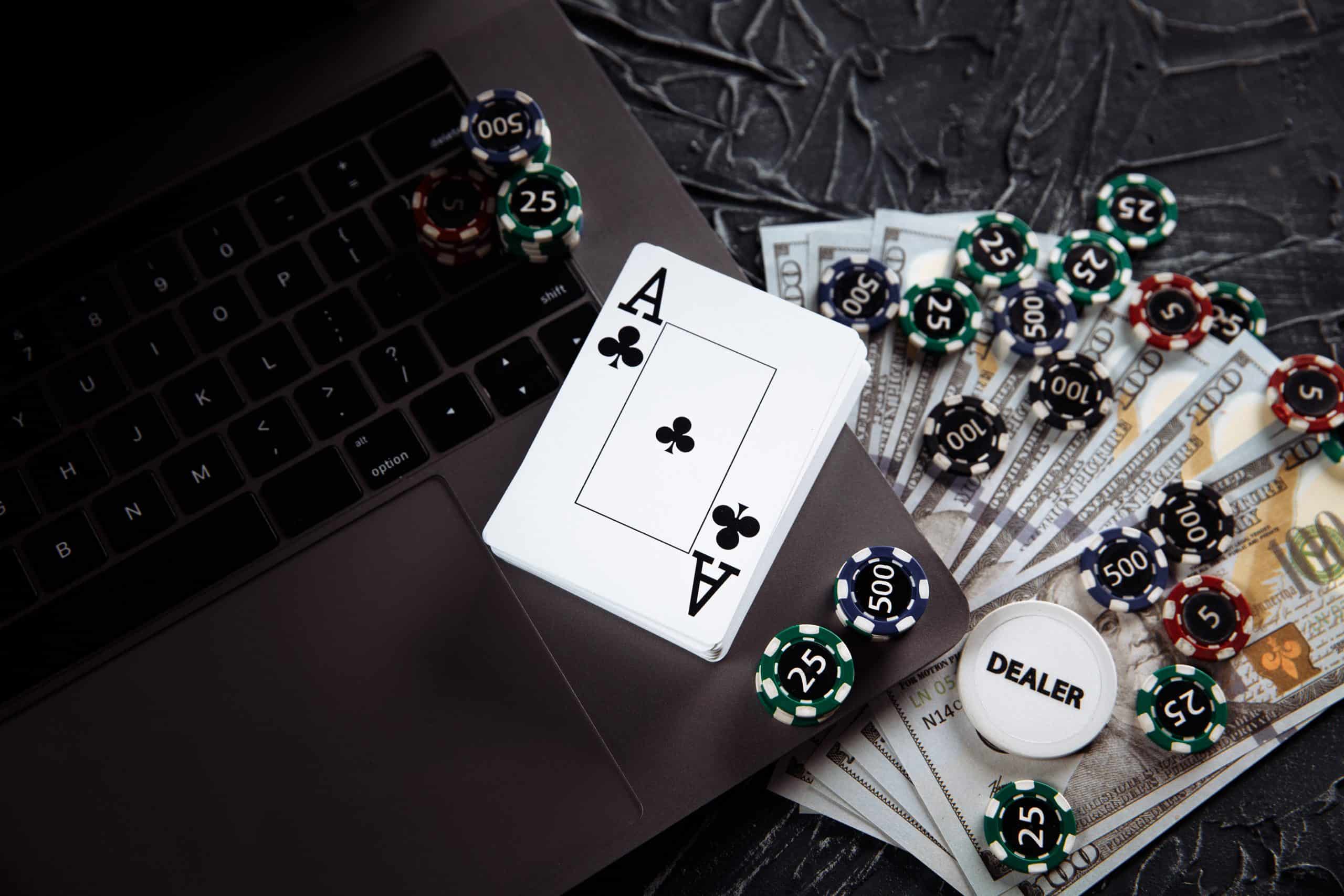 A laptop keyboard surrounded by casino chips, dollar bills, and playing cards, with an Ace of Clubs on top. A dealer button is also visible, suggesting an online gambling or poker game setting.