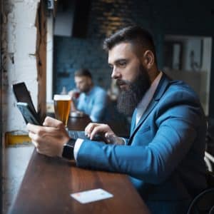 A man with a beard, wearing a blue suit, sits at a wooden counter in a cafe. He holds a smartphone and has a laptop open in front of him. A pint of beer is nearby, and another person is seated in the background.