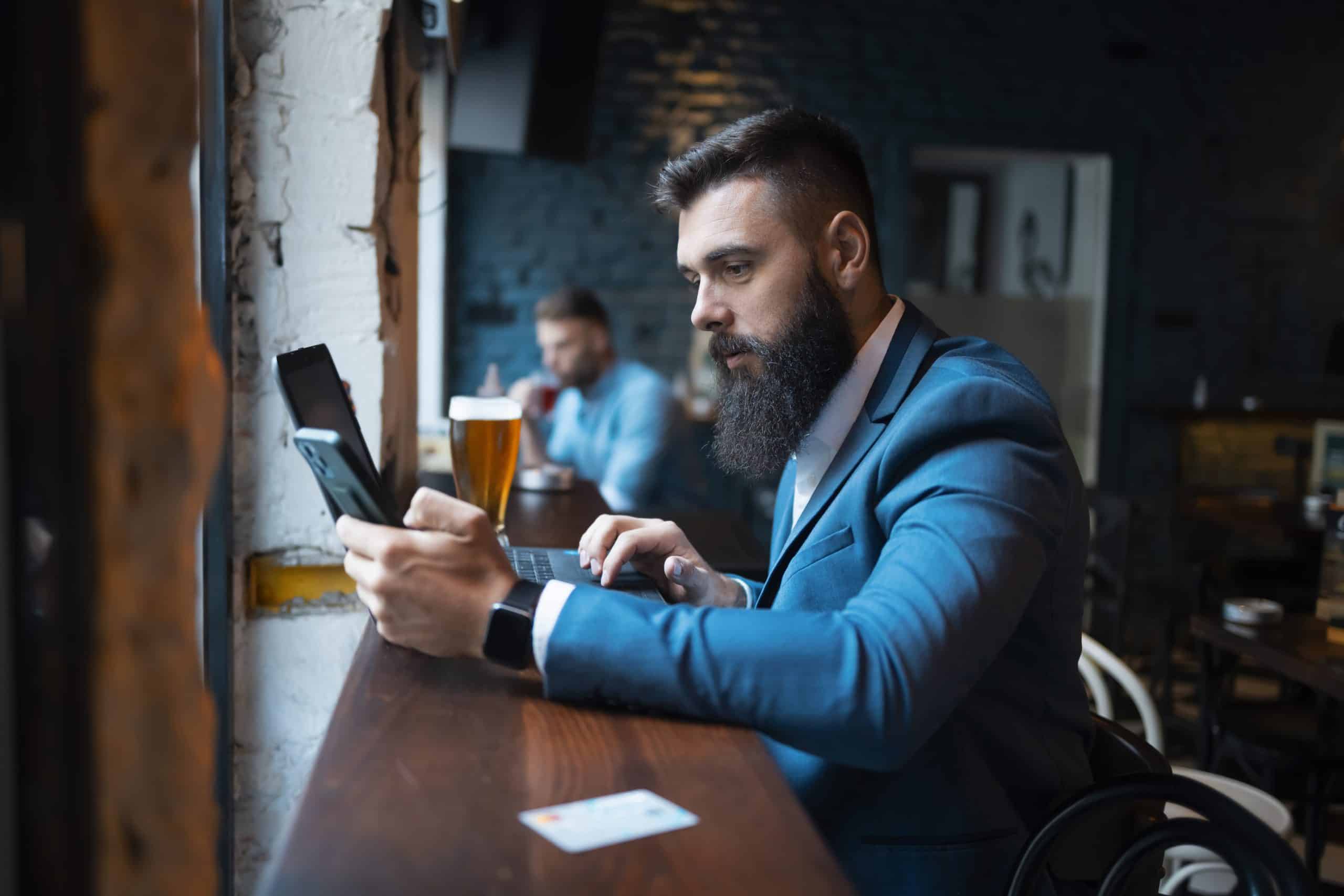 A man with a beard, wearing a blue suit, sits at a wooden counter in a cafe. He holds a smartphone and has a laptop open in front of him. A pint of beer is nearby, and another person is seated in the background.