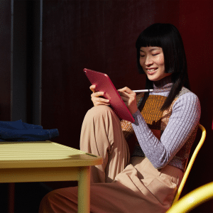 A person sitting at an outdoor table uses a stylus with a pink iPad 11. They're smiling, wearing a multicolored sweater and beige pants. The background features a red wall and an Apple logo in the corner.