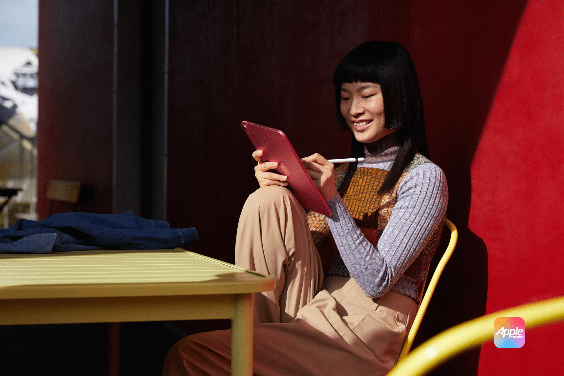 A person sitting at an outdoor table uses a stylus with a pink iPad 11. They're smiling, wearing a multicolored sweater and beige pants. The background features a red wall and an Apple logo in the corner.