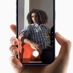 Close-up of a hand holding an iPhone 16, capturing a person seated elegantly on an orange chair. The phone screen displays the subject with camera settings visible. The white background and distinctive Apple logo hint at the recent sales surge of Apple's latest device.
