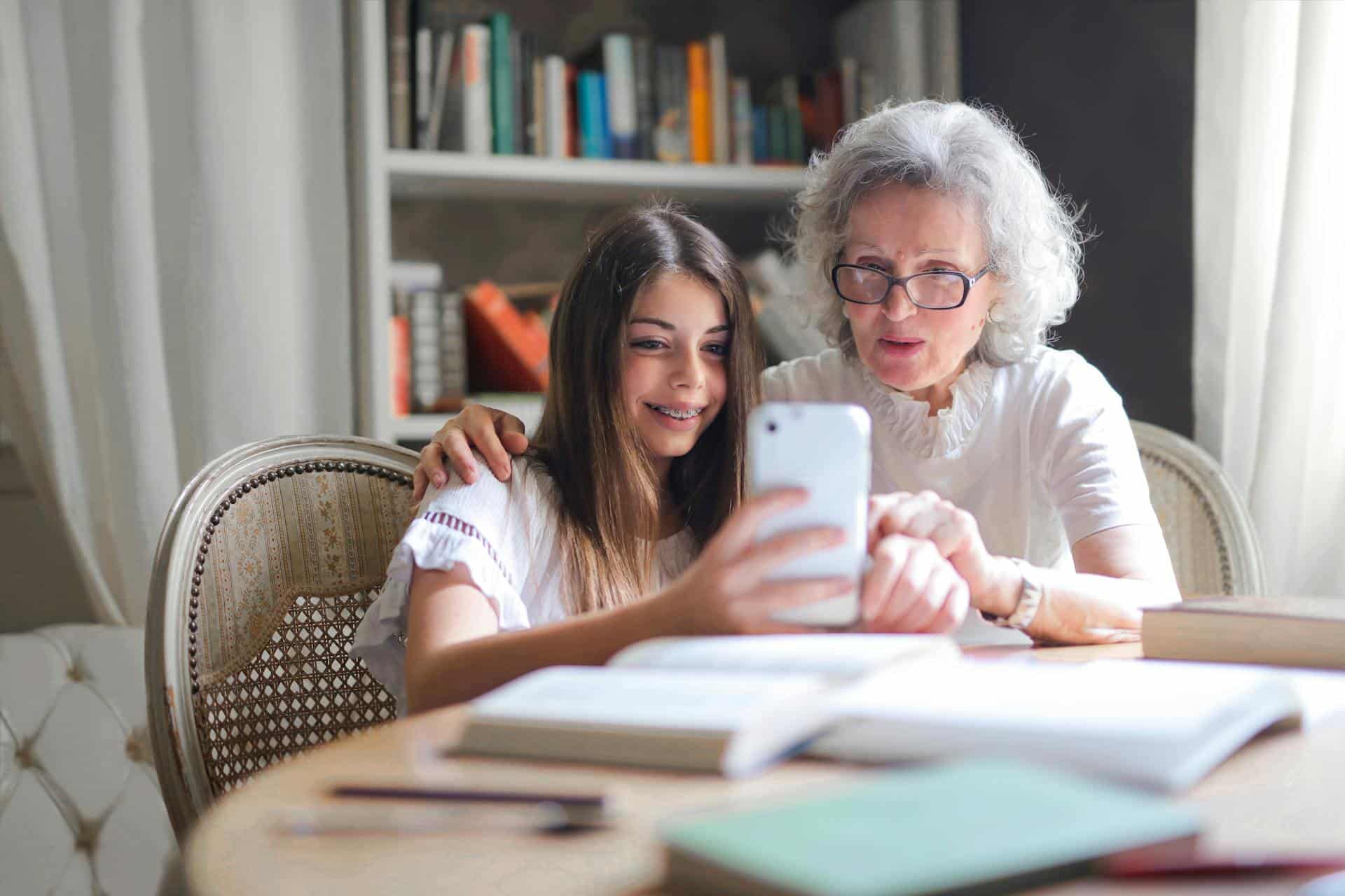 A young woman and an older woman sit together at a table, exploring the benefits of technology on a smartphone. The older woman, wearing glasses, smiles warmly as the young woman holds the phone. Books and a bookshelf fill the background, creating an inviting atmosphere for lifelong learning.