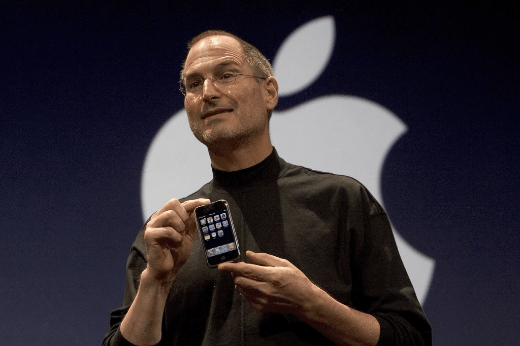 A person in a black turtleneck holds up an iPhone in front of a large, partially visible logo. The dark background emphasizes the sleek device and the logo.