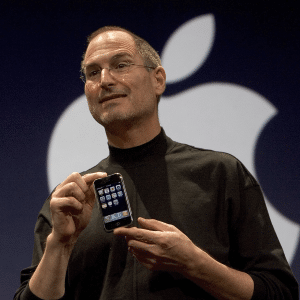 A person in a black turtleneck holds up an iPhone in front of a large, partially visible logo. The dark background emphasizes the sleek device and the logo.