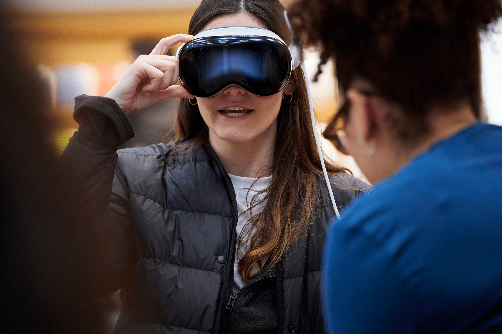 A person wearing a virtual reality headset, possibly a lower-priced Apple Vision Headset, interacts with augmented reality. Another person, slightly blurred, faces them, perhaps assisting or discussing the technology indoors.