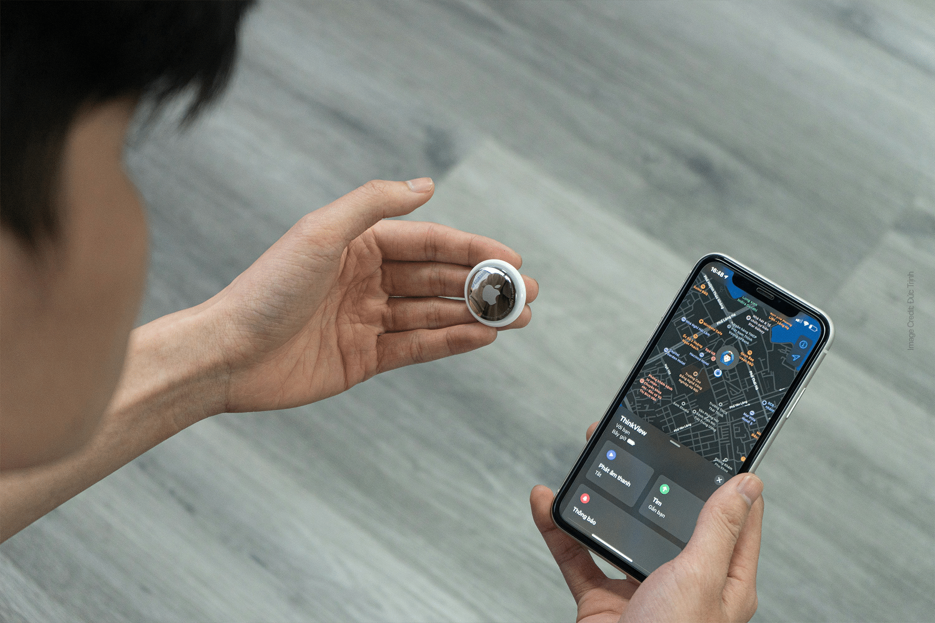 A person holding an American Airlines AirTag in their left hand and an iPhone in their right, displaying a map with location details on the screen. The background is a light wood floor, providing a warm and inviting ambiance to this tech-savvy moment.