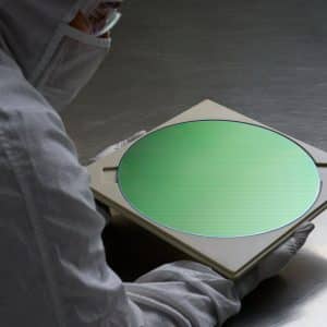 A person in a cleanroom suit and gloves holds a green silicon wafer on a beige tray, possibly for use in AI servers. The setting is a sterile lab environment, with a metallic surface gleaming in the background.