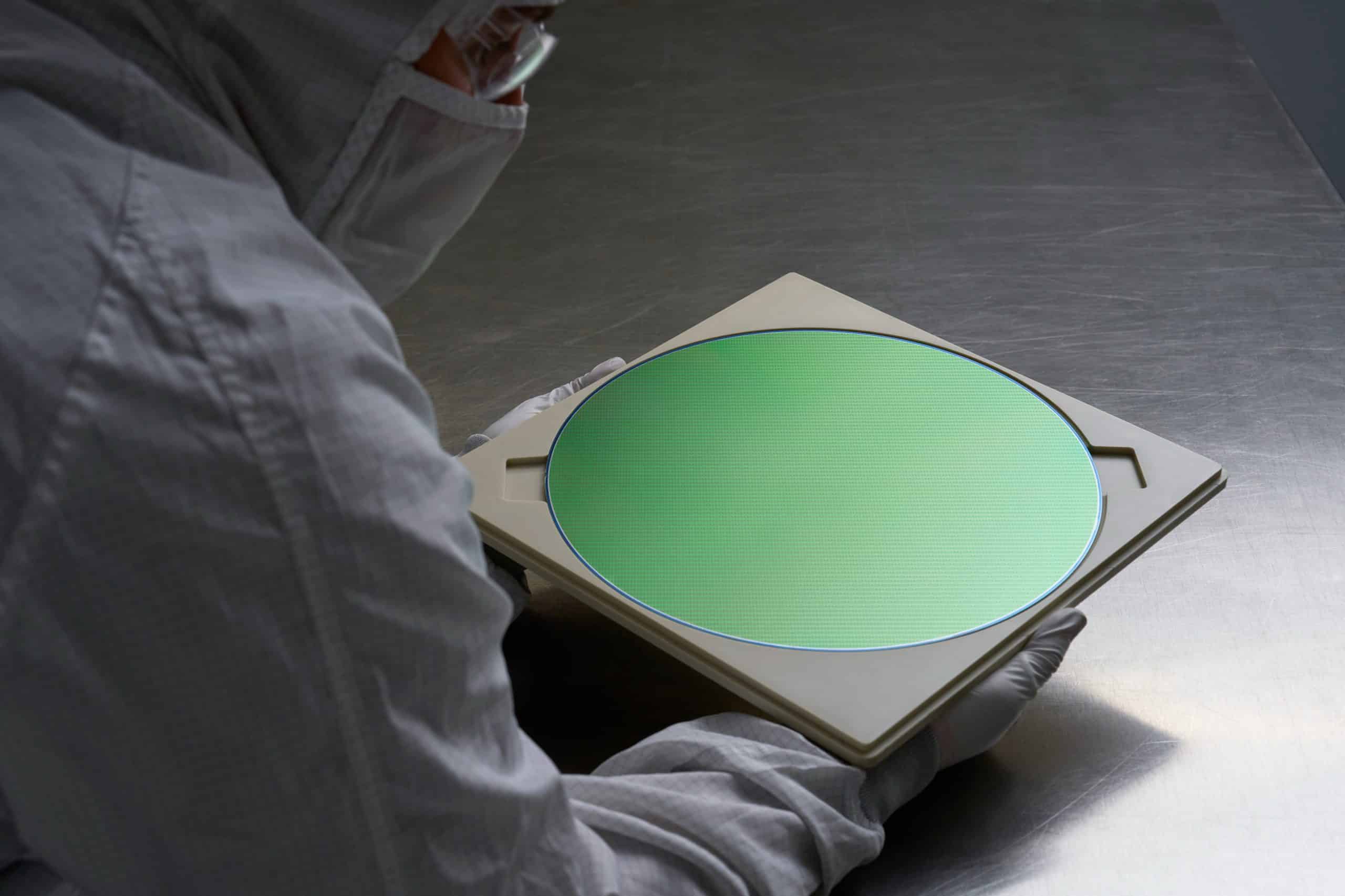 A person in a cleanroom suit and gloves holds a green silicon wafer on a beige tray, possibly for use in AI servers. The setting is a sterile lab environment, with a metallic surface gleaming in the background.