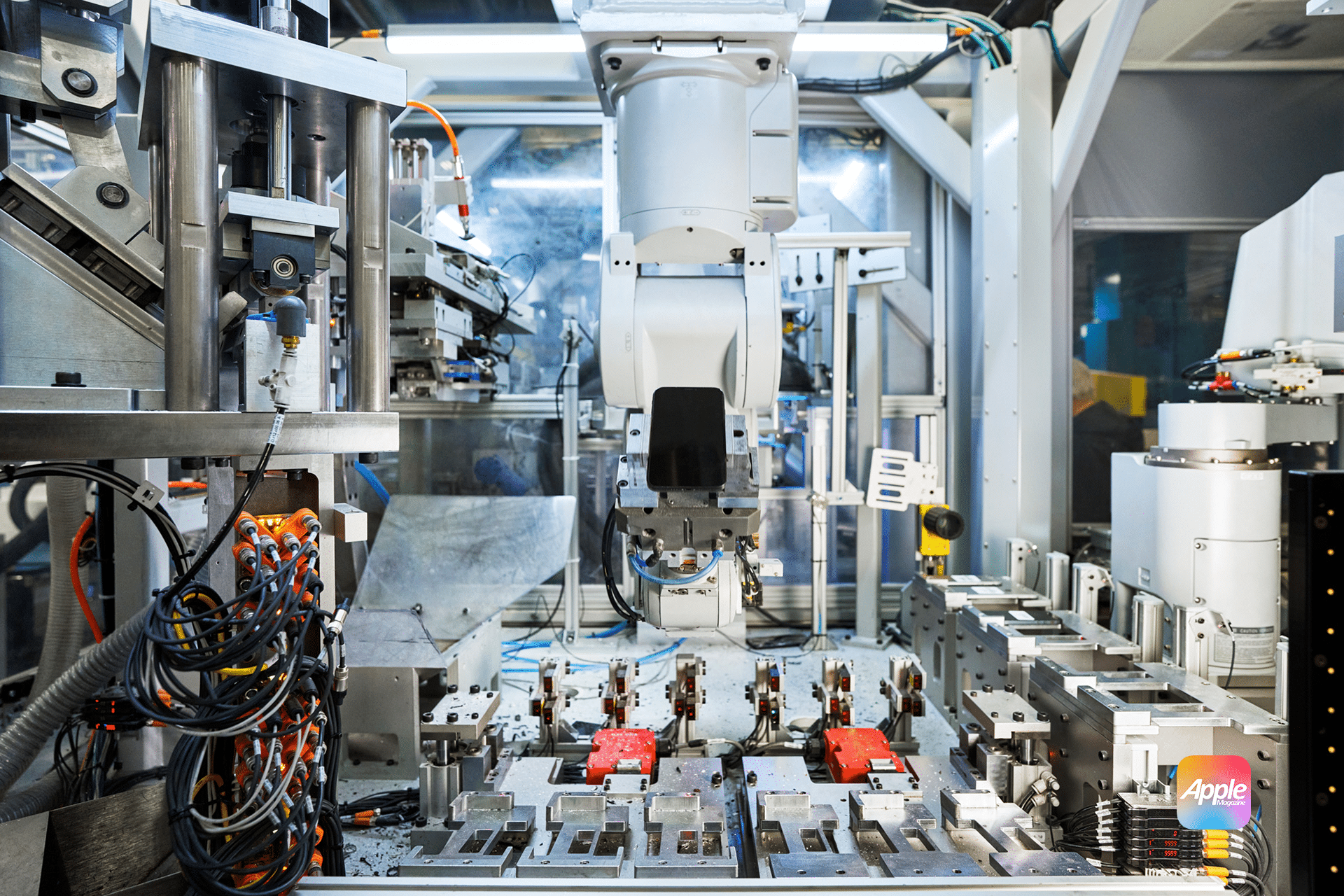 Inside a high-tech manufacturing facility, an Apple robot engages in assembling electronic components. The workspace is filled with machinery, wiring, and several red components on a platform. A subtle glow illuminates the area below a small logo featuring “Apple” in the corner.