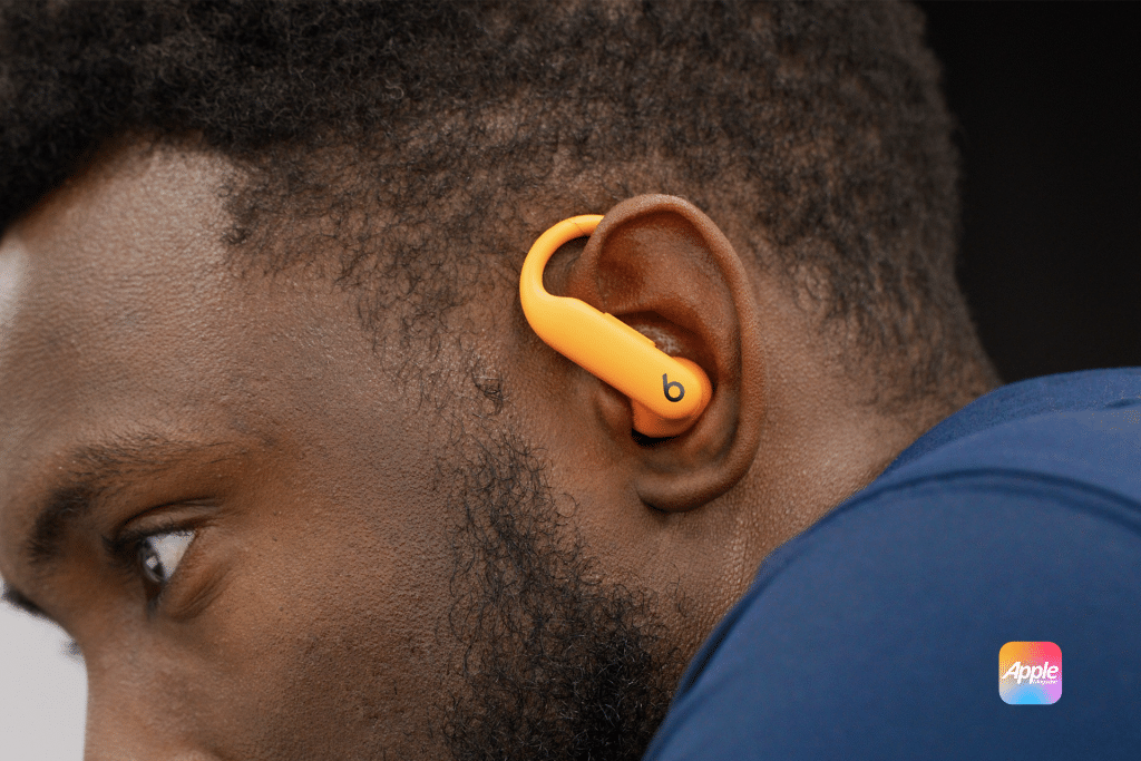 Close-up of a person wearing an orange Powerbeats Pro 2 earbud in their left ear. The individual, with short hair and a navy blue top, showcases the visible Beats logo on the earbud, while a small Apple logo sits in the bottom right corner.