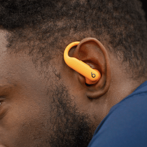 Close-up of a person wearing an orange Powerbeats Pro 2 earbud in their left ear. The individual, with short hair and a navy blue top, showcases the visible Beats logo on the earbud, while a small Apple logo sits in the bottom right corner.