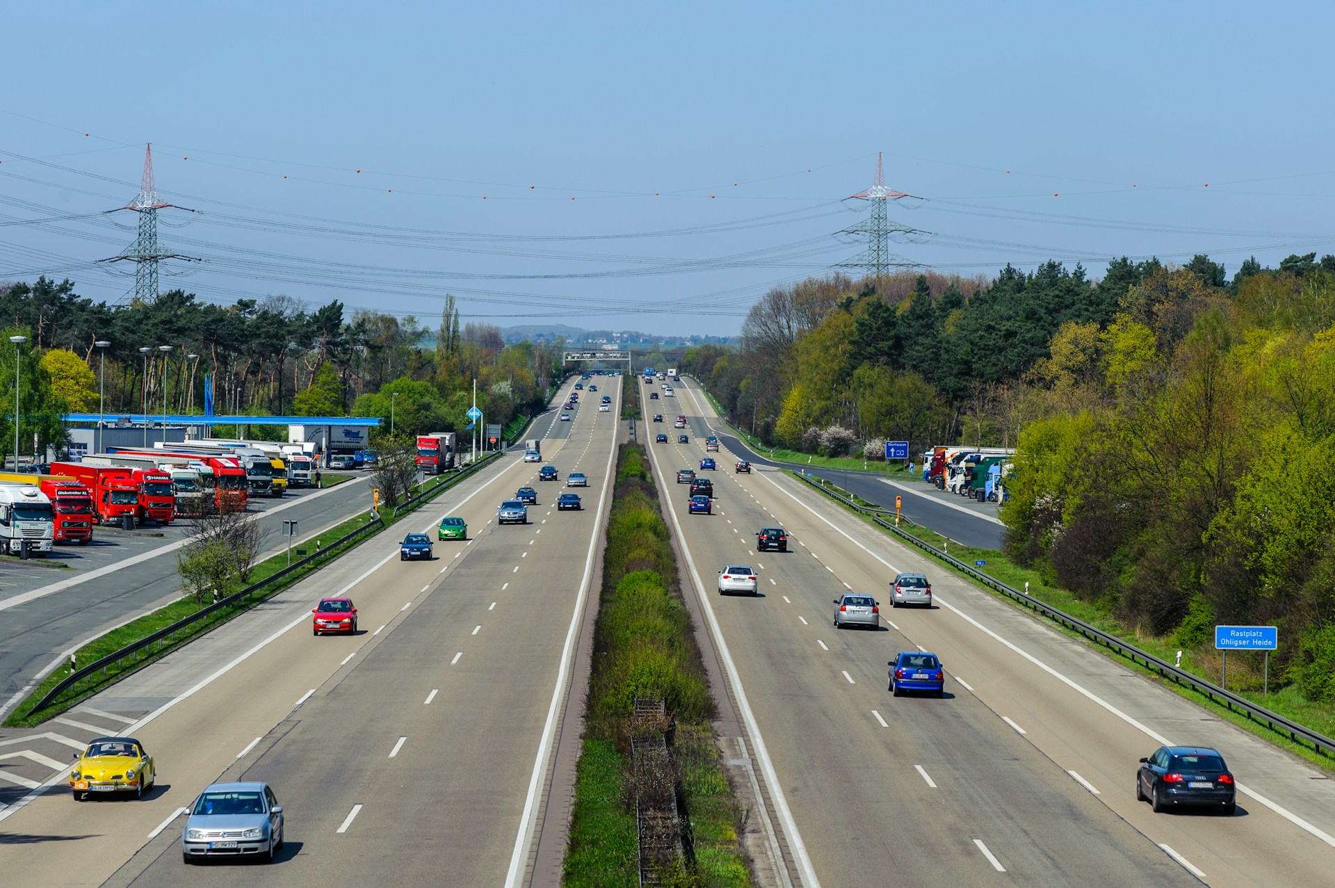 An aerial view shows a busy highway bustling with cars and trucks, perhaps some heading to review a first settlement offer after a car accident. Lush greenery lines the sides as power lines cross above. On the left, a rest stop hosts parked trucks under the sprawling sky.