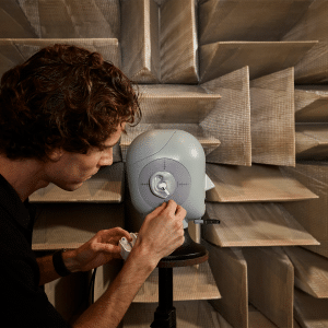 A person adjusts a small speaker on a stand in a soundproof room with acoustic panels. The geometric-patterned walls enhance sound absorption, creating an ideal environment for testing audio equipment, perhaps exploring the AirPods Pro 2's hearing aid feature with focused precision.