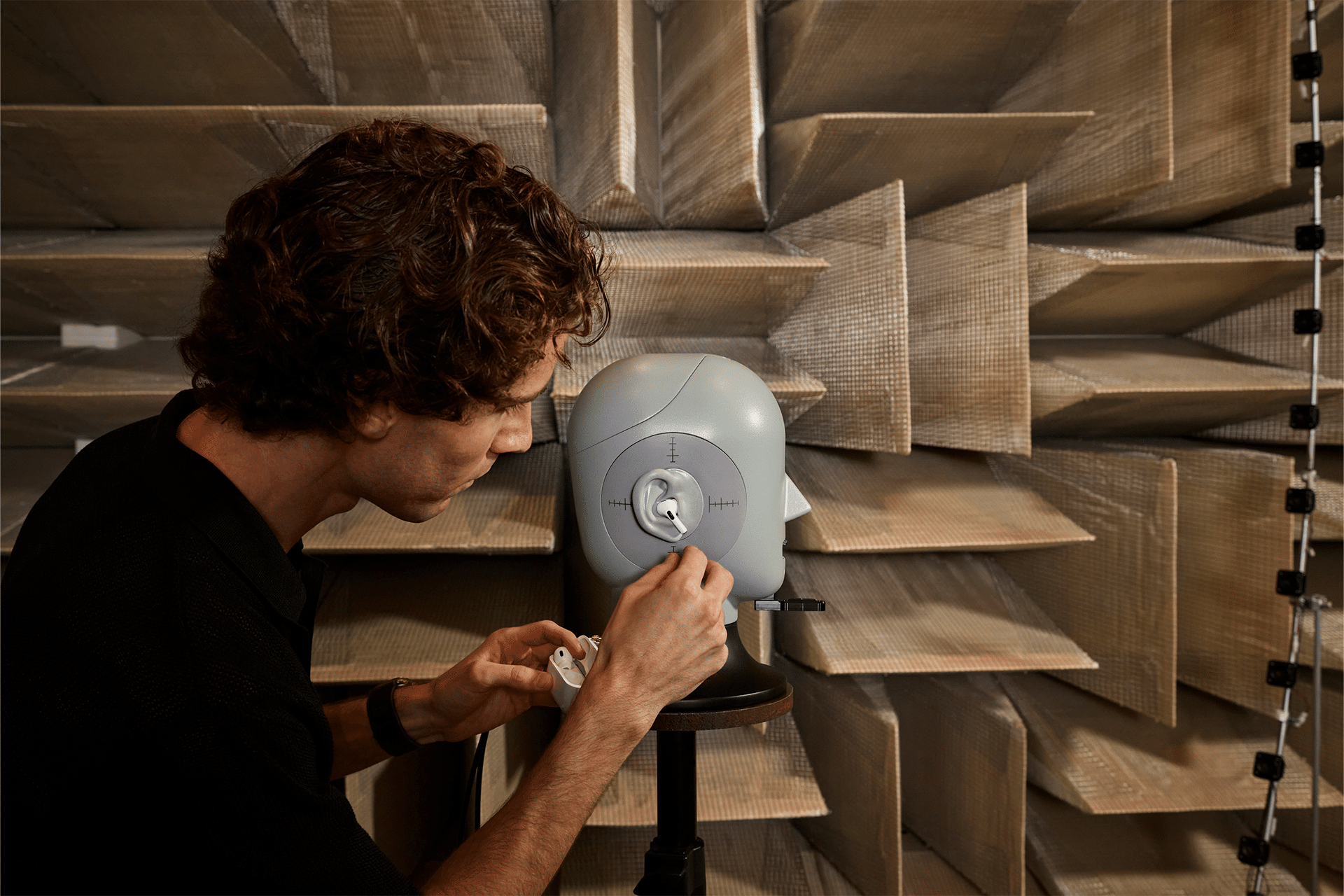 A person adjusts a small speaker on a stand in a soundproof room with acoustic panels. The geometric-patterned walls enhance sound absorption, creating an ideal environment for testing audio equipment, perhaps exploring the AirPods Pro 2's hearing aid feature with focused precision.