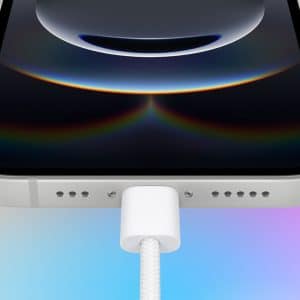 A smartphone with a silver edge is shown from the bottom, with a white charging cable plugged into it. The screen displays a dark circular design. The background is a gradient of orange, pink, and blue. An Apple logo is in the lower right corner.