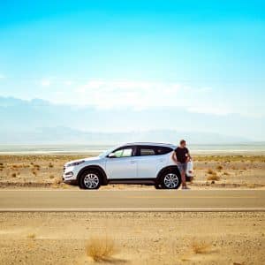 A person stands beside a parked white SUV on a deserted road in a vast, arid desert landscape, reminiscent of Earth's first settlements. The sky is clear and blue, with distant mountains on the horizon. Sparse, dry vegetation dots the sandy ground.