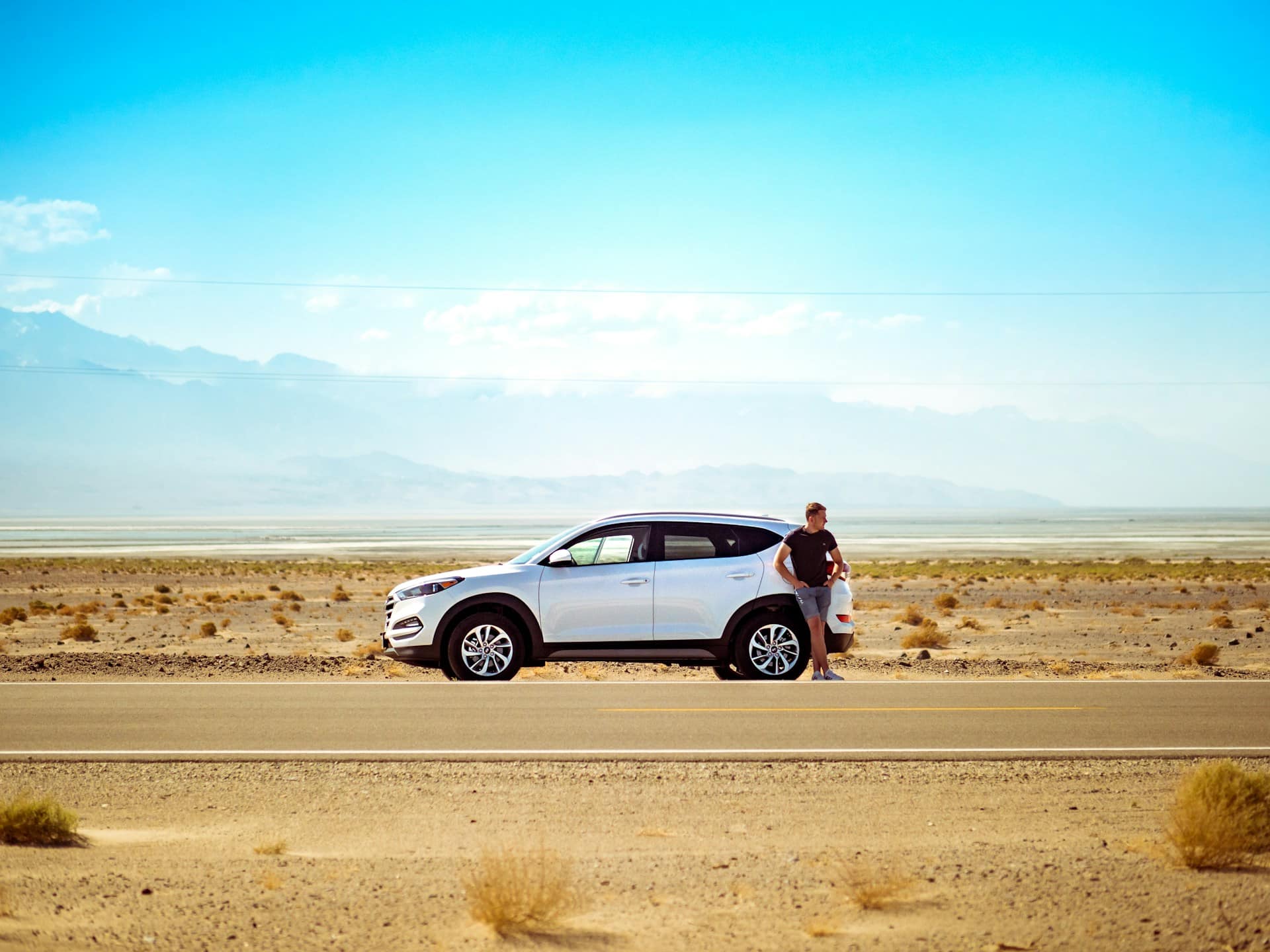 A person stands beside a parked white SUV on a deserted road in a vast, arid desert landscape, reminiscent of Earth's first settlements. The sky is clear and blue, with distant mountains on the horizon. Sparse, dry vegetation dots the sandy ground.