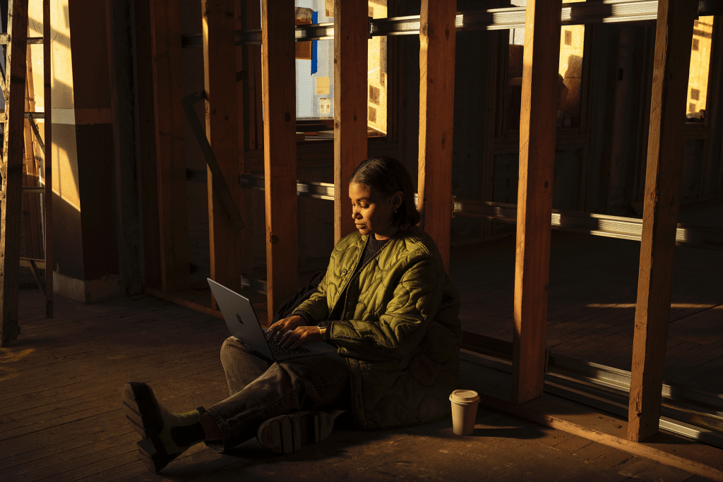 A person in a green jacket sits on the floor against a wooden beam, using a MacBook Pro with an M5 Chip. A coffee cup is nearby, while warm light from a window creates a cozy atmosphere in the partially constructed or renovated space.