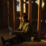 A person in a green jacket sits on the floor against a wooden beam, using a MacBook Pro with an M5 Chip. A coffee cup is nearby, while warm light from a window creates a cozy atmosphere in the partially constructed or renovated space.