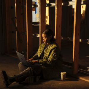 A person in a green jacket sits on the floor against a wooden beam, using a MacBook Pro with an M5 Chip. A coffee cup is nearby, while warm light from a window creates a cozy atmosphere in the partially constructed or renovated space.