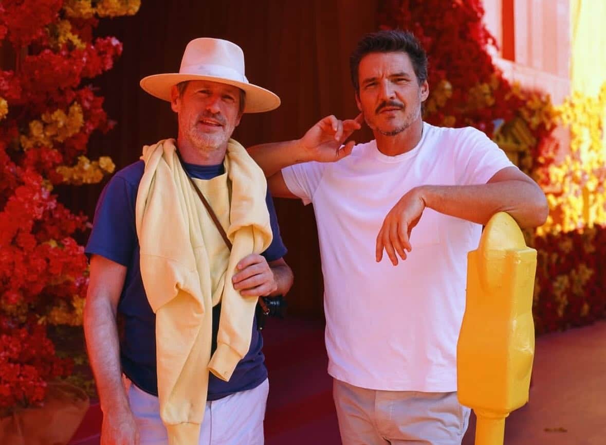 Two men pose in front of vibrant red and yellow floral decorations. One wears a wide-brimmed hat, dark shirt, and a yellow sweater draped over his shoulders like Pedro Pascal might. The other, seemingly lost in thought with AirPods 4 shimmering, rests an arm on a yellow object.