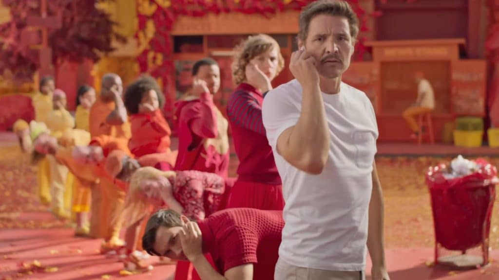 A man in a white T-shirt, wearing AirPods 4, gestures towards his eye as he stands before a line of people in colorful outfits. The vibrant background features shades of red and orange, with autumn leaves scattered on the ground.