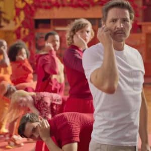 A man in a white T-shirt, wearing AirPods 4, gestures towards his eye as he stands before a line of people in colorful outfits. The vibrant background features shades of red and orange, with autumn leaves scattered on the ground.