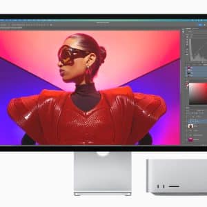 A Mac Studio monitor displays an image editing software interface, featuring a person in futuristic red attire and sunglasses against a vibrant pink and purple background. A silver computer tower is shown beneath the screen, highlighting sleek design and advanced capabilities.