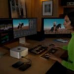 A person in a neon green shirt sits at a desk with a Mac Studio, editing video and color on a multi-monitor setup. The workspace features external drives, control panels, photos on the wall, and a shelf filled with books.